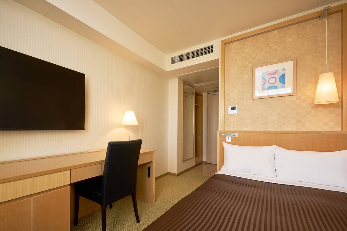 A hotel room with a bed, a desk with a chair, a television, a lamp, and a door leading to another room.
