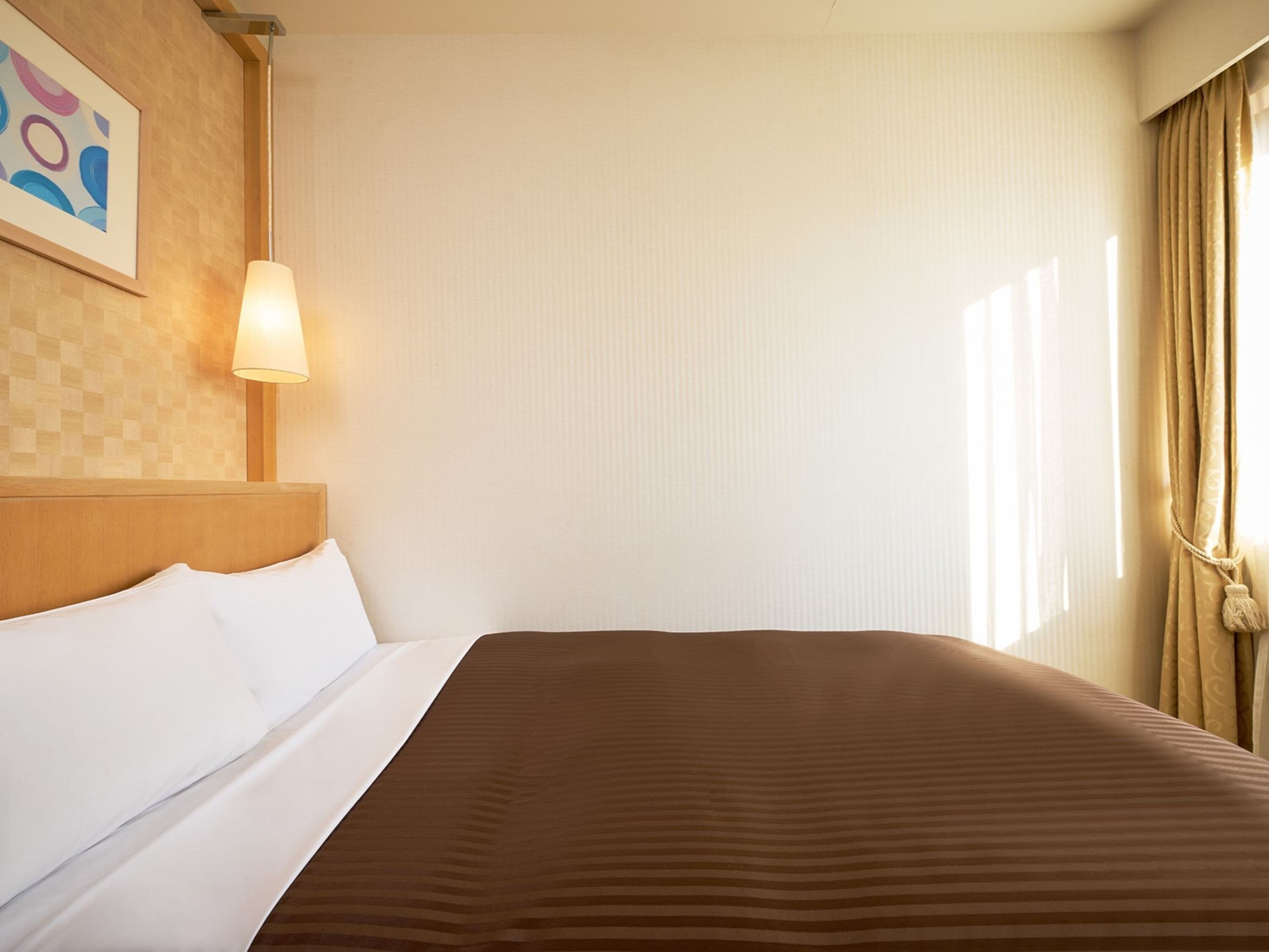 A hotel room with a bed, a headboard, a wall-mounted lamp, and a window with curtains.