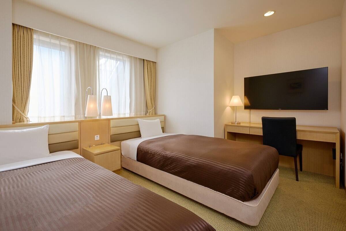 A hotel room with two beds, a desk, a TV, and a window with curtains.