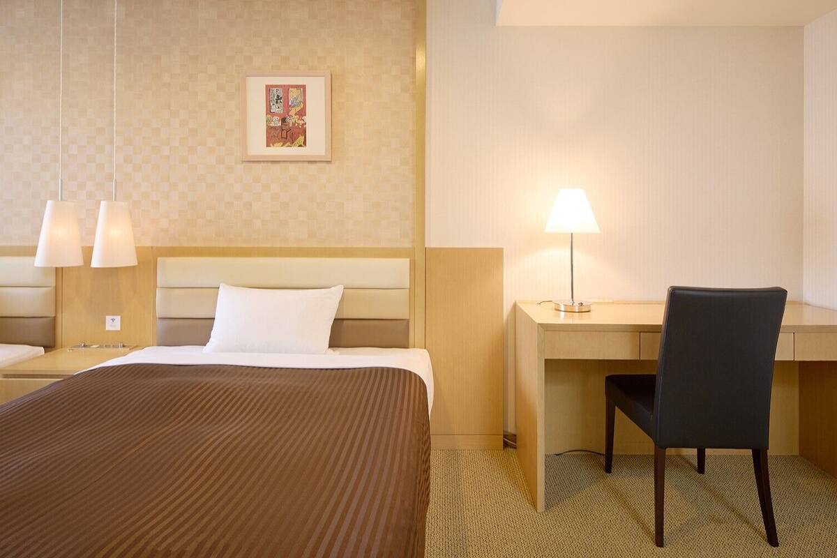 A hotel room with two beds, a desk, a chair, and two wall-mounted lamps.