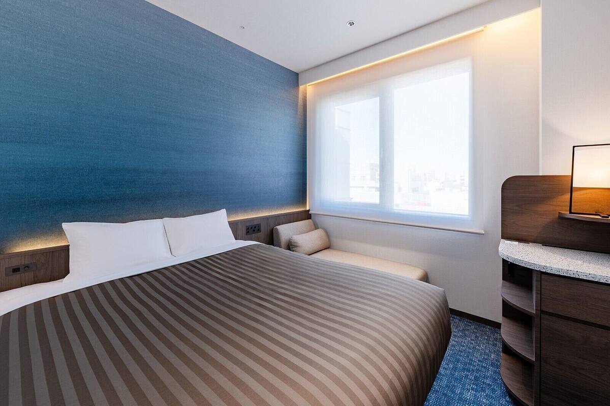 A hotel room with a large bed, a window with a view, a desk with a lamp, and a blue accent wall.