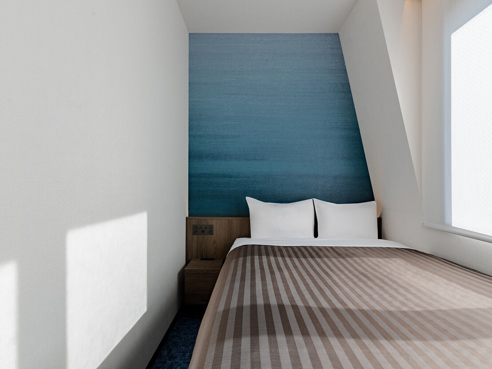 Une chambre avec un plafond en pente, un lit recouvert d’une couverture à rayures et une fenêtre sur le côté droit.