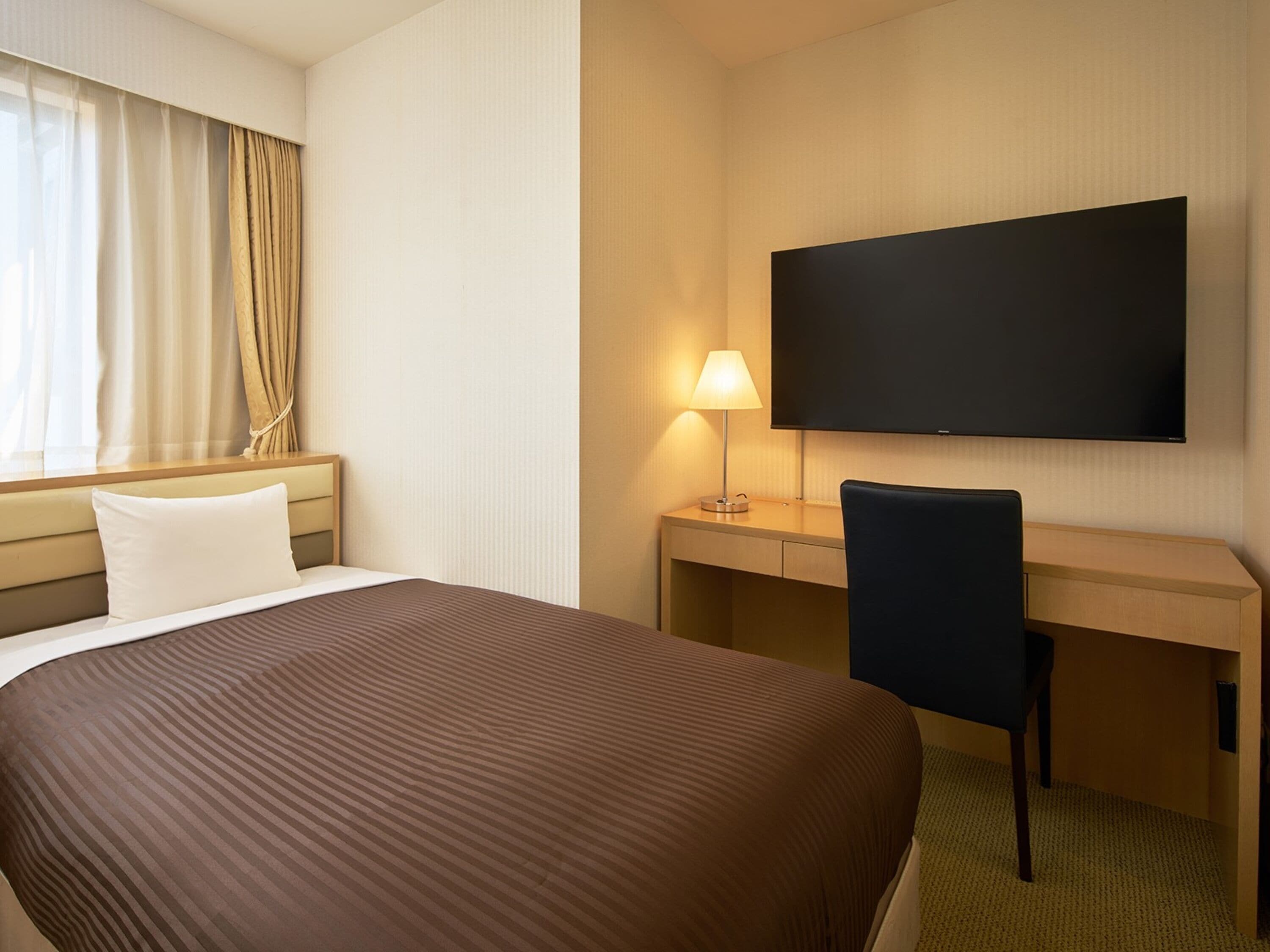 A hotel room with a large bed, a desk with a chair, a flat-screen TV, and a lamp.