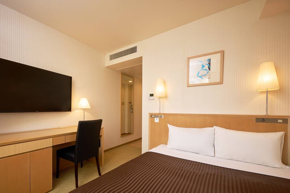 A hotel room with a bed, a desk, a chair, a TV, and two bedside lamps.