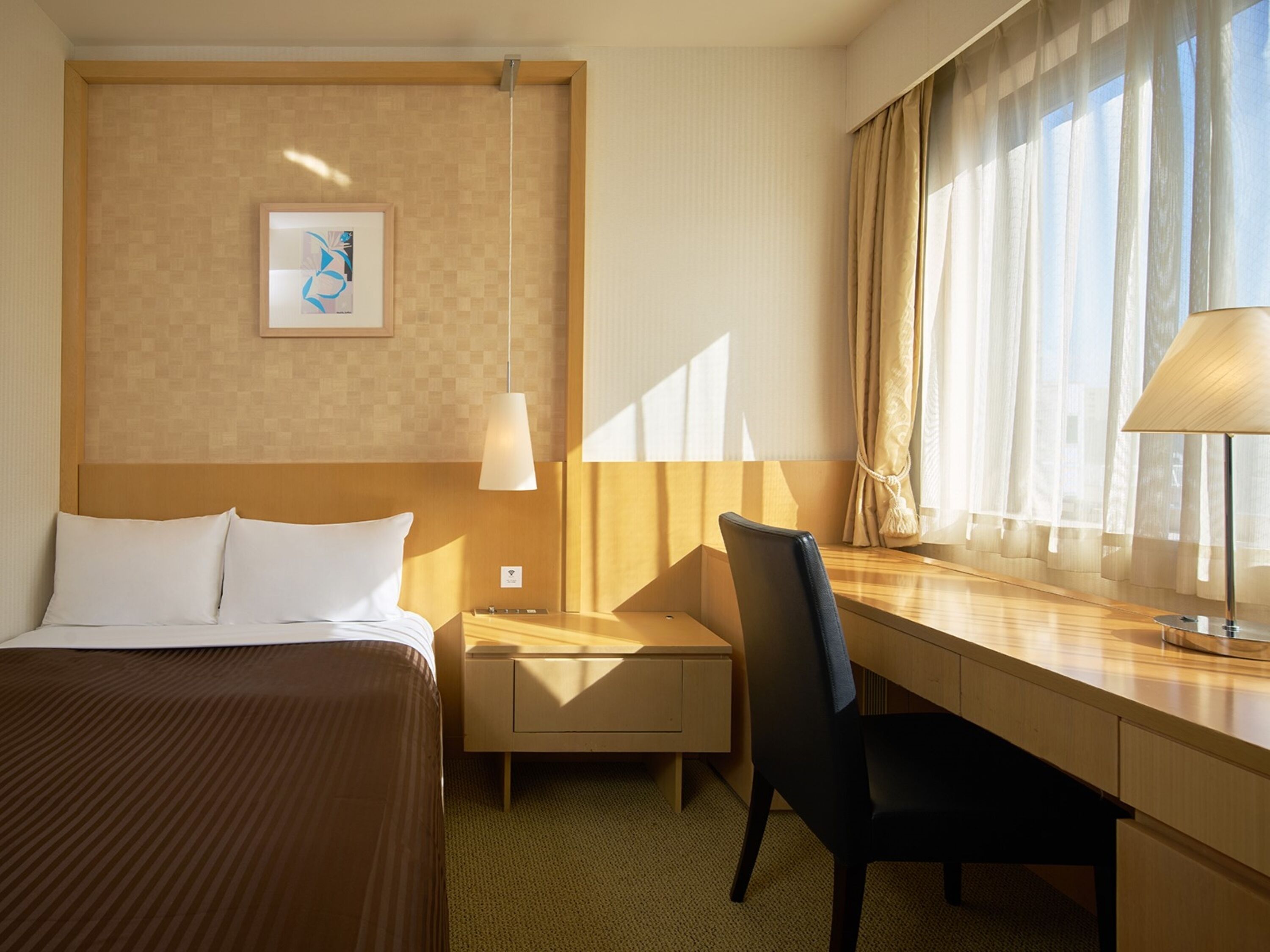 A hotel room with a bed, a desk, a chair, a lamp, a window with curtains, and a framed picture on the wall.
