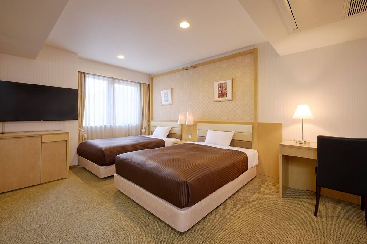 A hotel room with two beds, a desk, a chair, a TV, and a window with curtains.