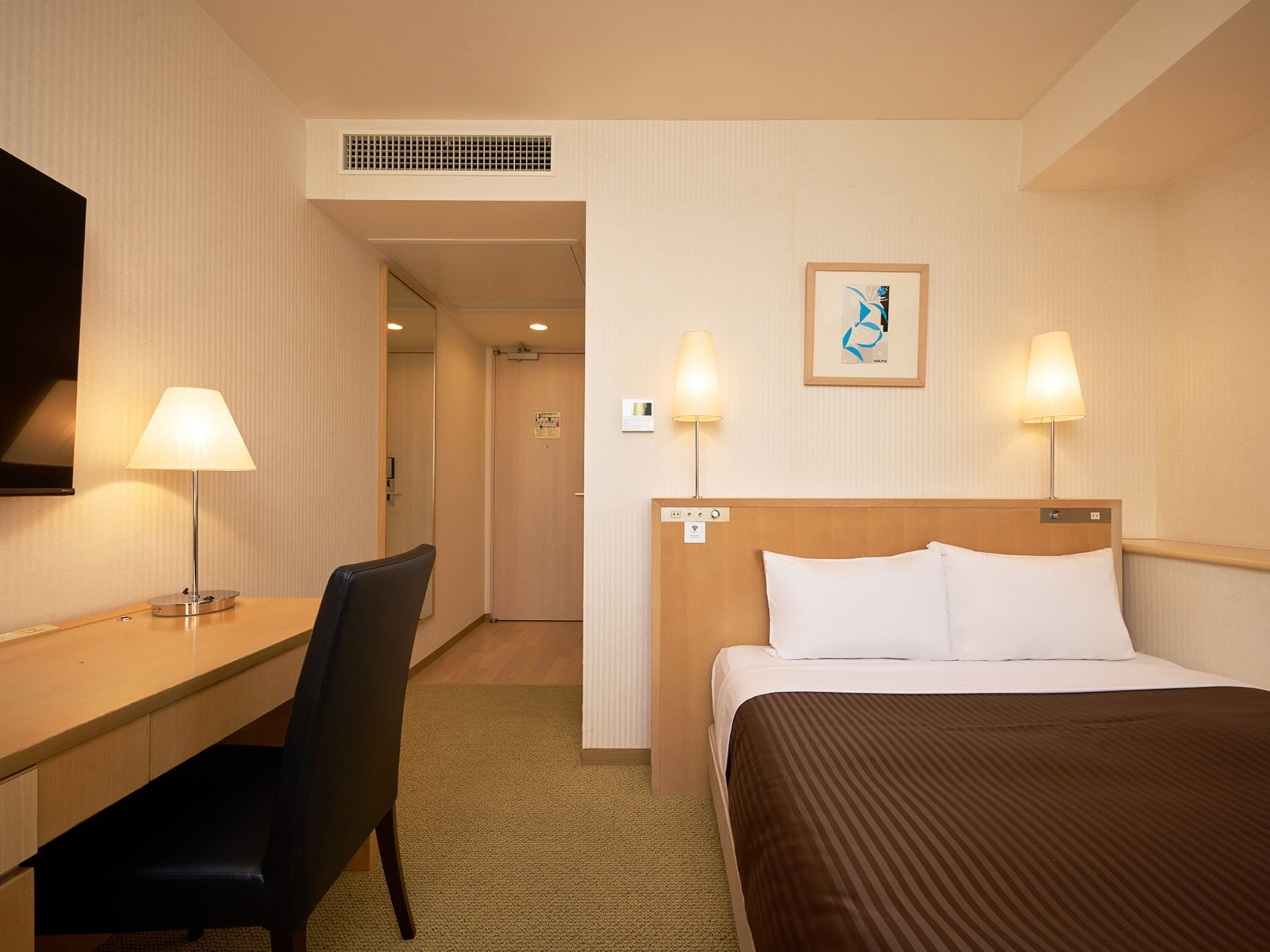 Ett hotellrum med en säng, ett skrivbord, en stol, två lampor, en tv och en inramad tavla på väggen.