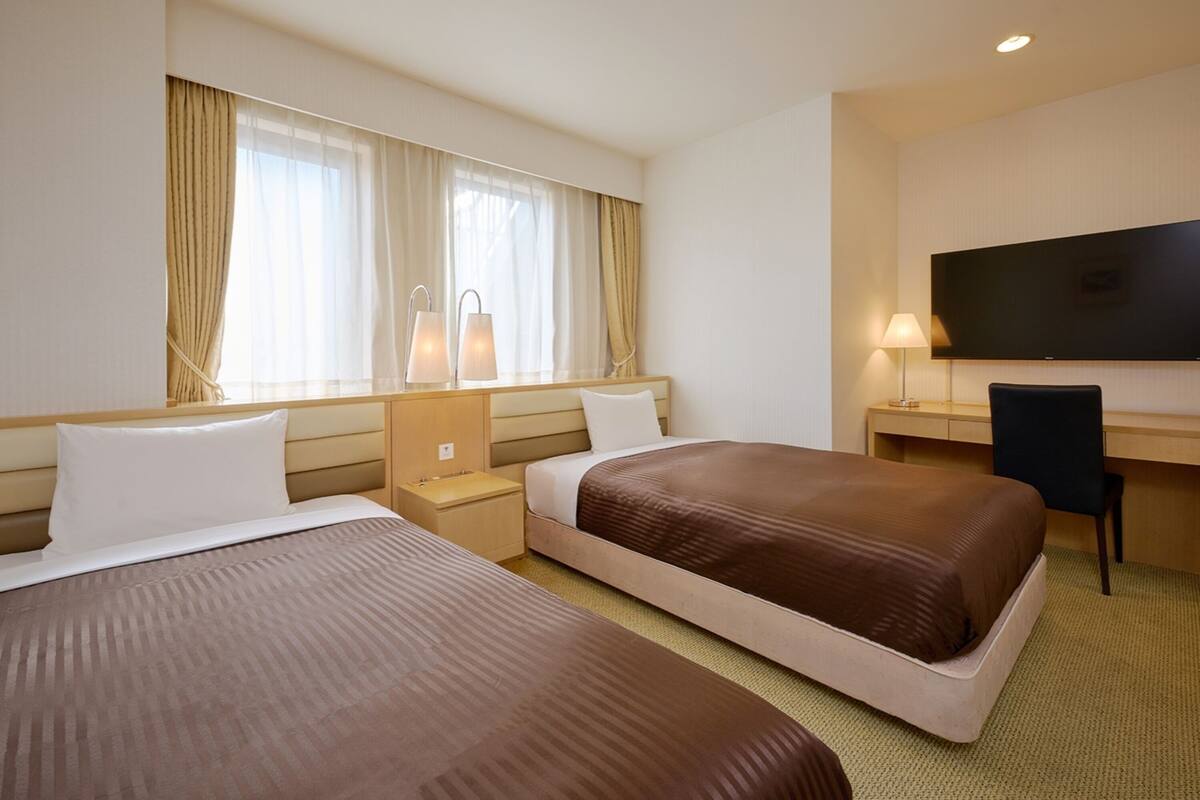 A hotel room with two beds, a desk, a TV, and a window with curtains.