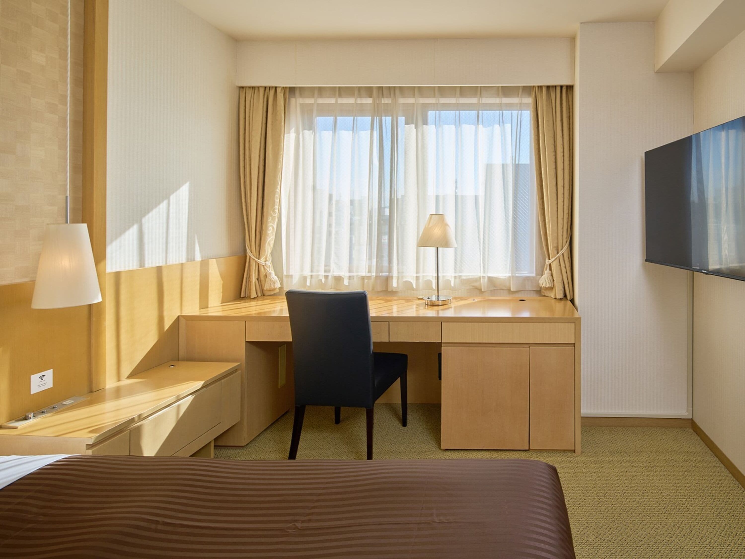 A hotel room with a bed, a desk with a chair, a lamp, and a TV on the wall.