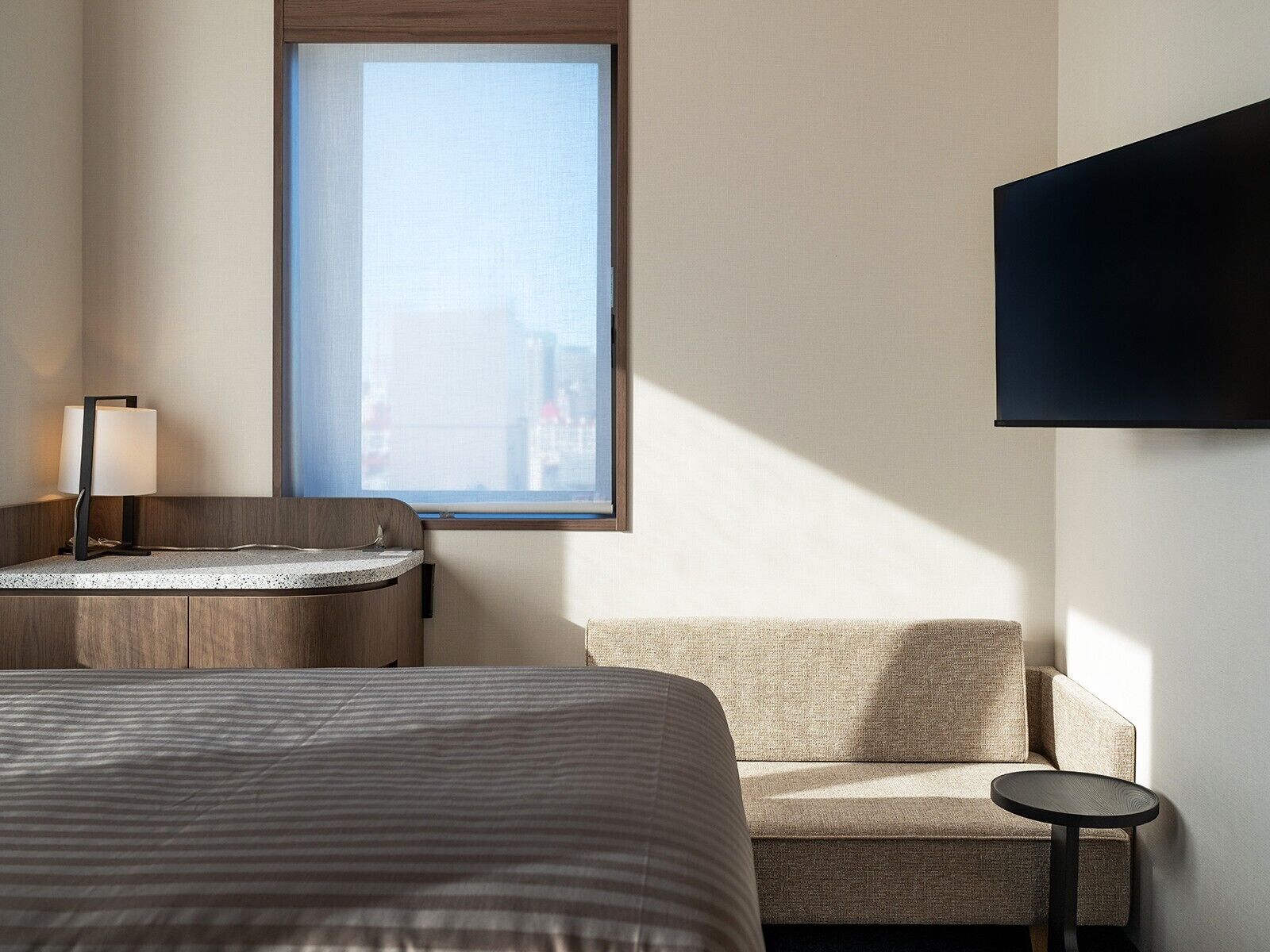 A hotel room with a bed, a small round table, a sofa, a desk with a lamp, and a TV mounted on the wall.