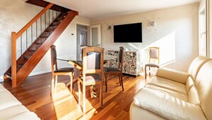 Een woonkamer met een houten trap, een eethoek met stoelen, een bank en een flatscreen televisie aan de muur.