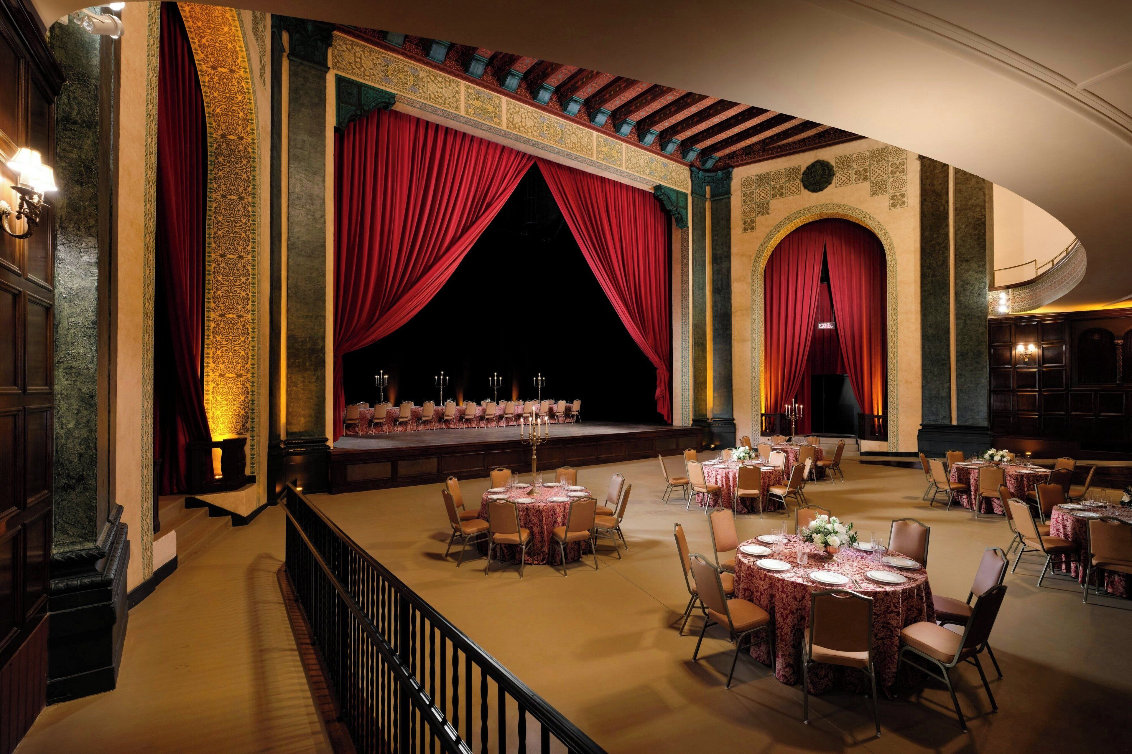 A grand hall with a stage, red curtains, and multiple round tables with chairs.