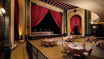 A grand hall with a stage, red curtains, and multiple round tables with chairs.