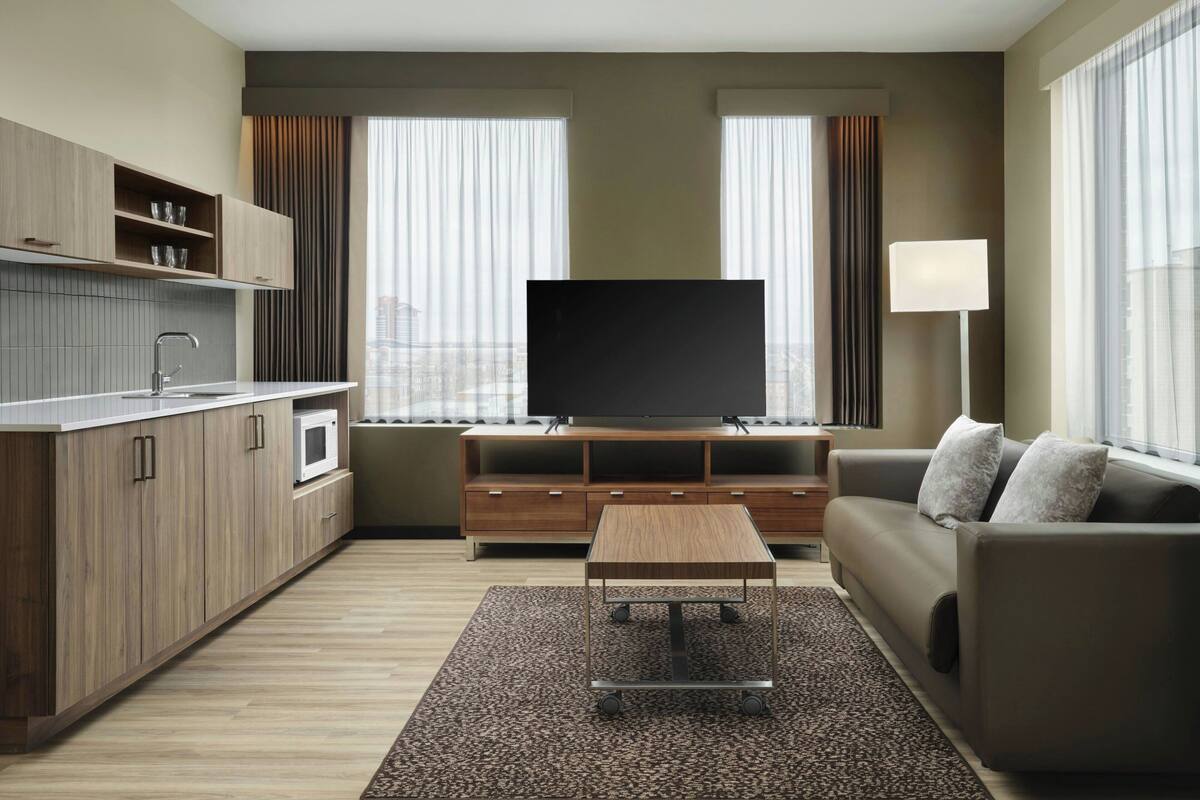 A modern hotel room with a kitchenette, a flat-screen TV, a sofa, and a large window with curtains.