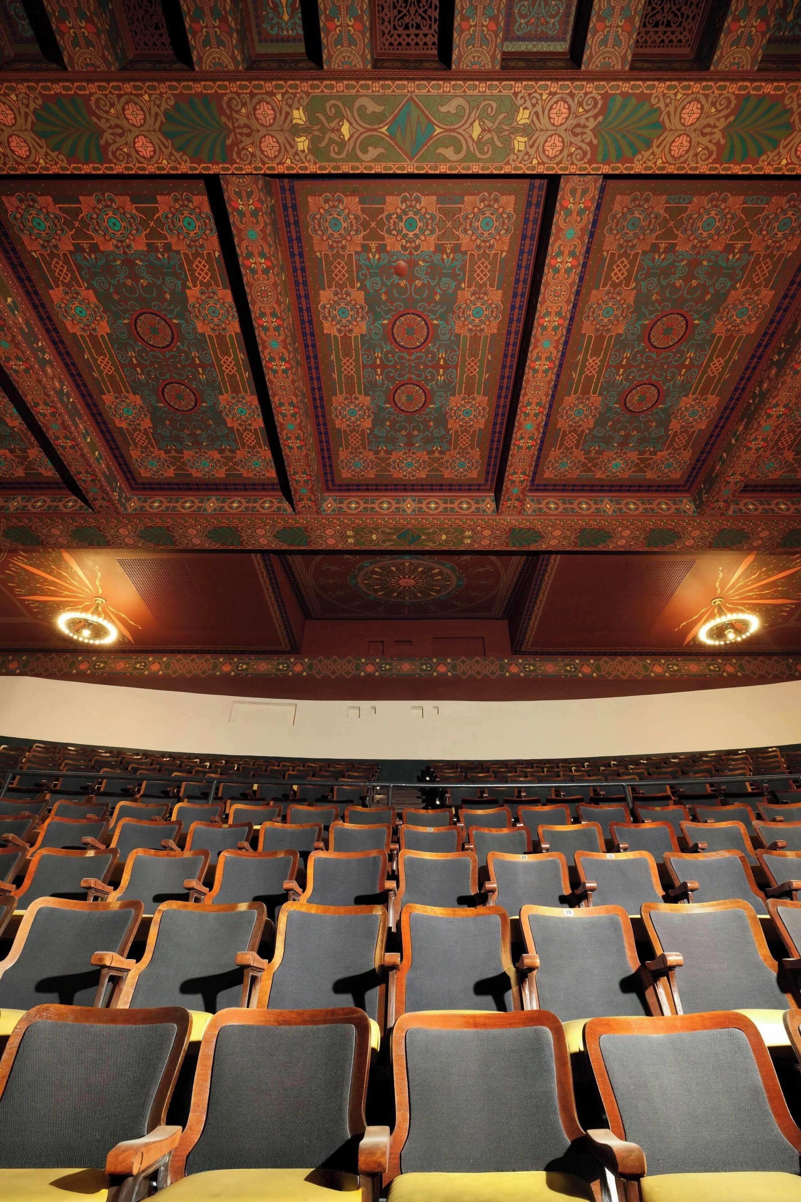 A theater with ornate ceiling patterns and rows of wooden chairs.