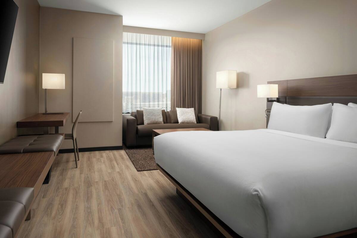 A modern hotel room with a large bed, a sofa, a desk, and a view of the city through large windows.