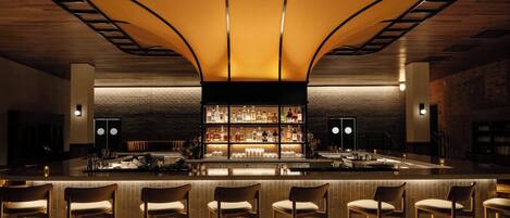 A bar area with a central island, bar stools, and a well-lit shelf displaying bottles.
