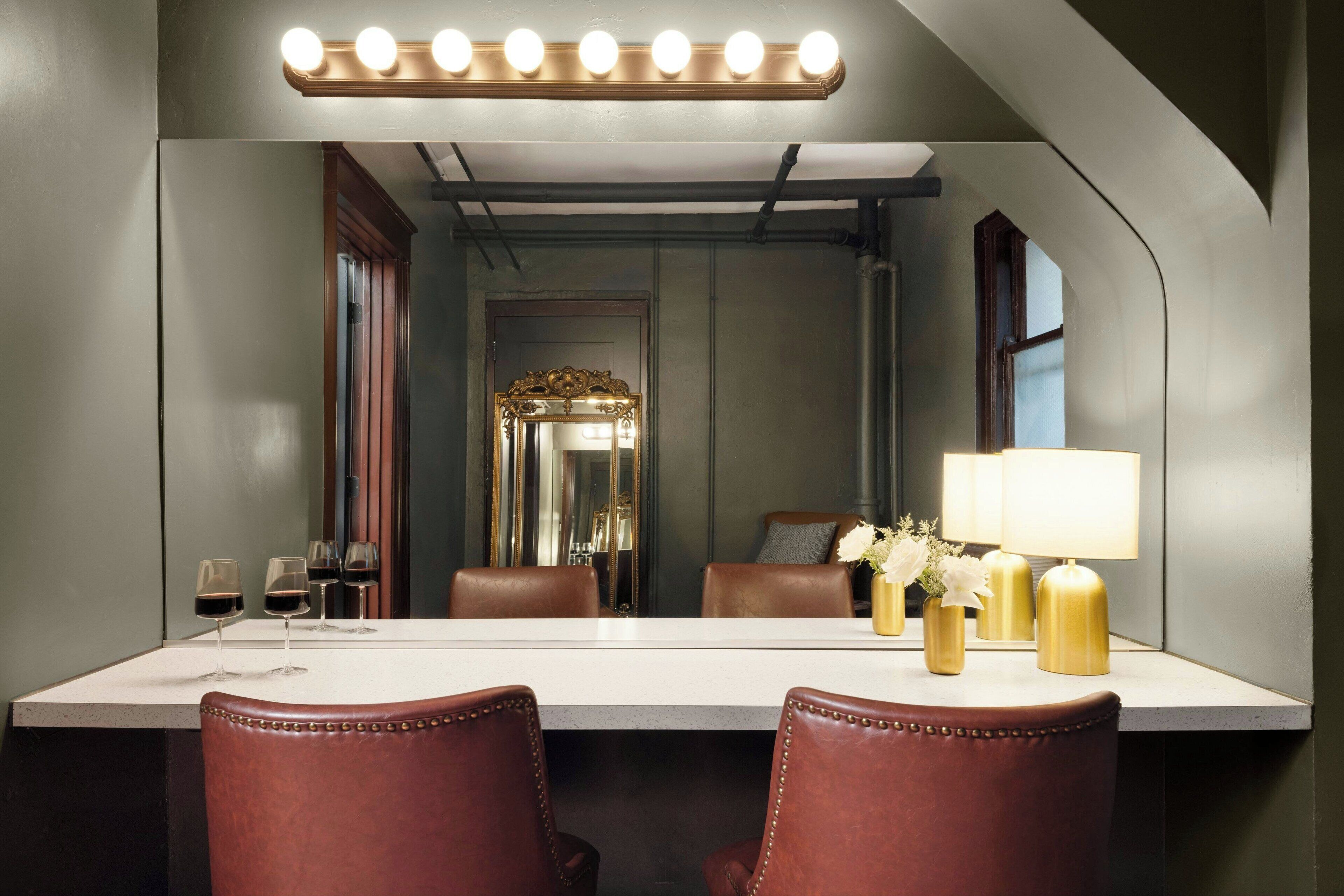 A hotel room with a large mirror, a vanity table with a lamp, and a seating area with leather chairs.