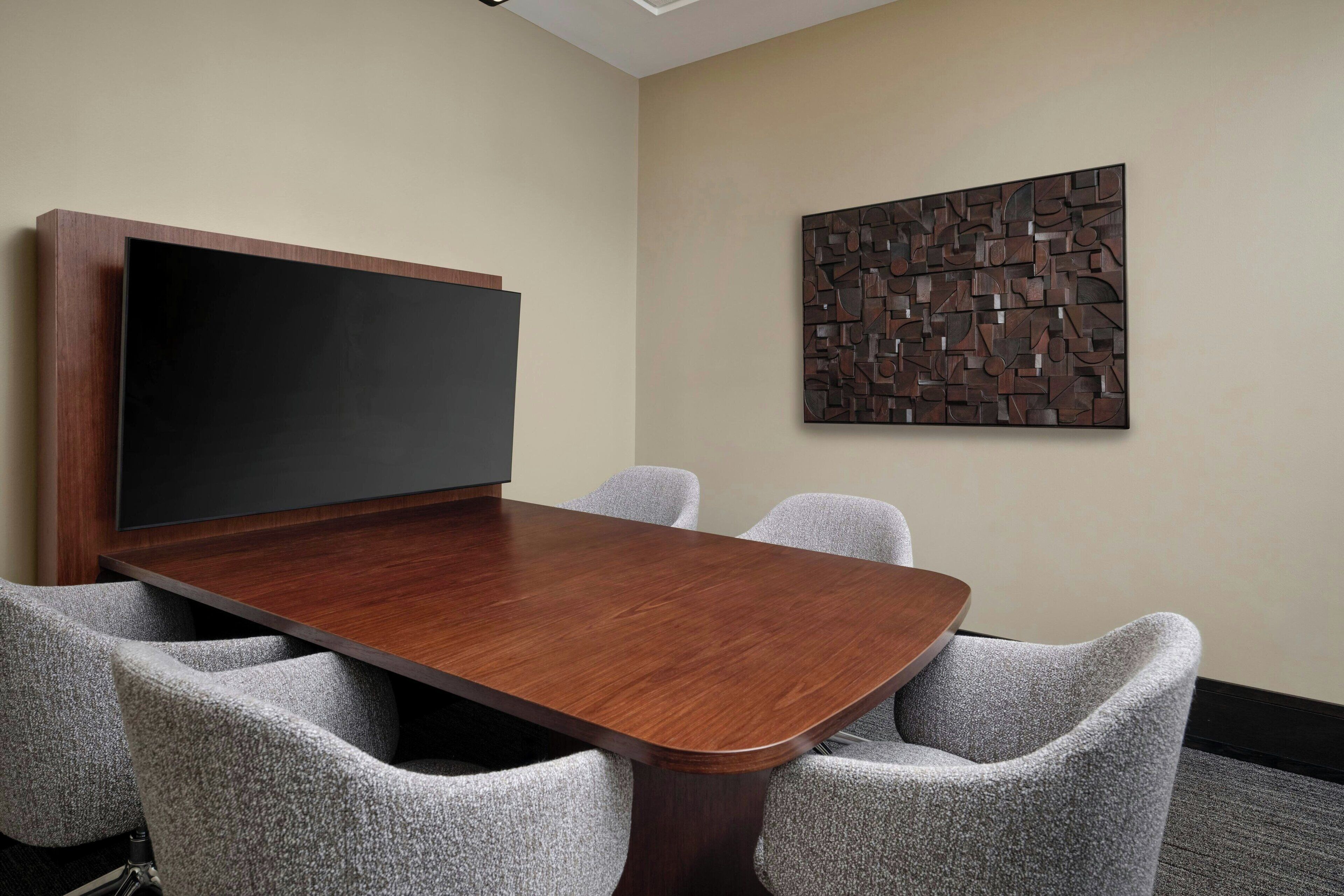 A conference room with a wooden table, grey upholstered chairs, a large flat-screen TV, and a geometric wall art piece.