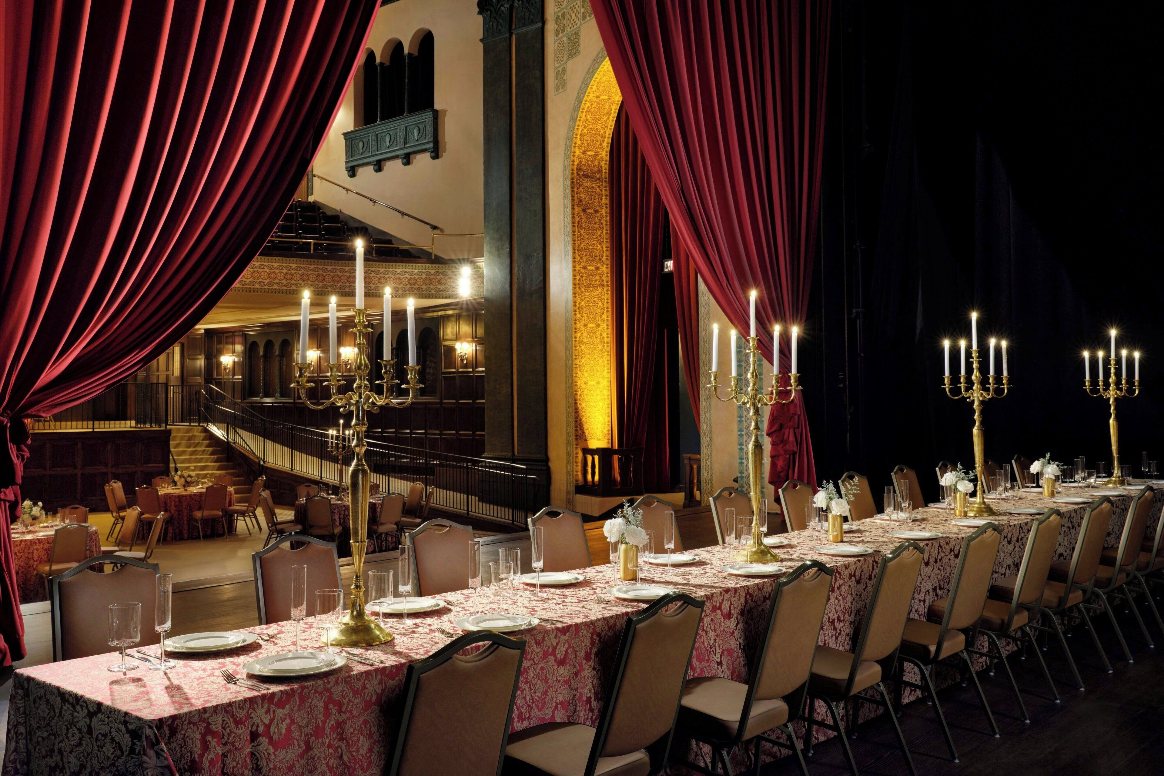 Une salle à manger élégante, avec une longue table dressée pour un repas, des rideaux rouges et des chandeliers illuminés à la bougie.