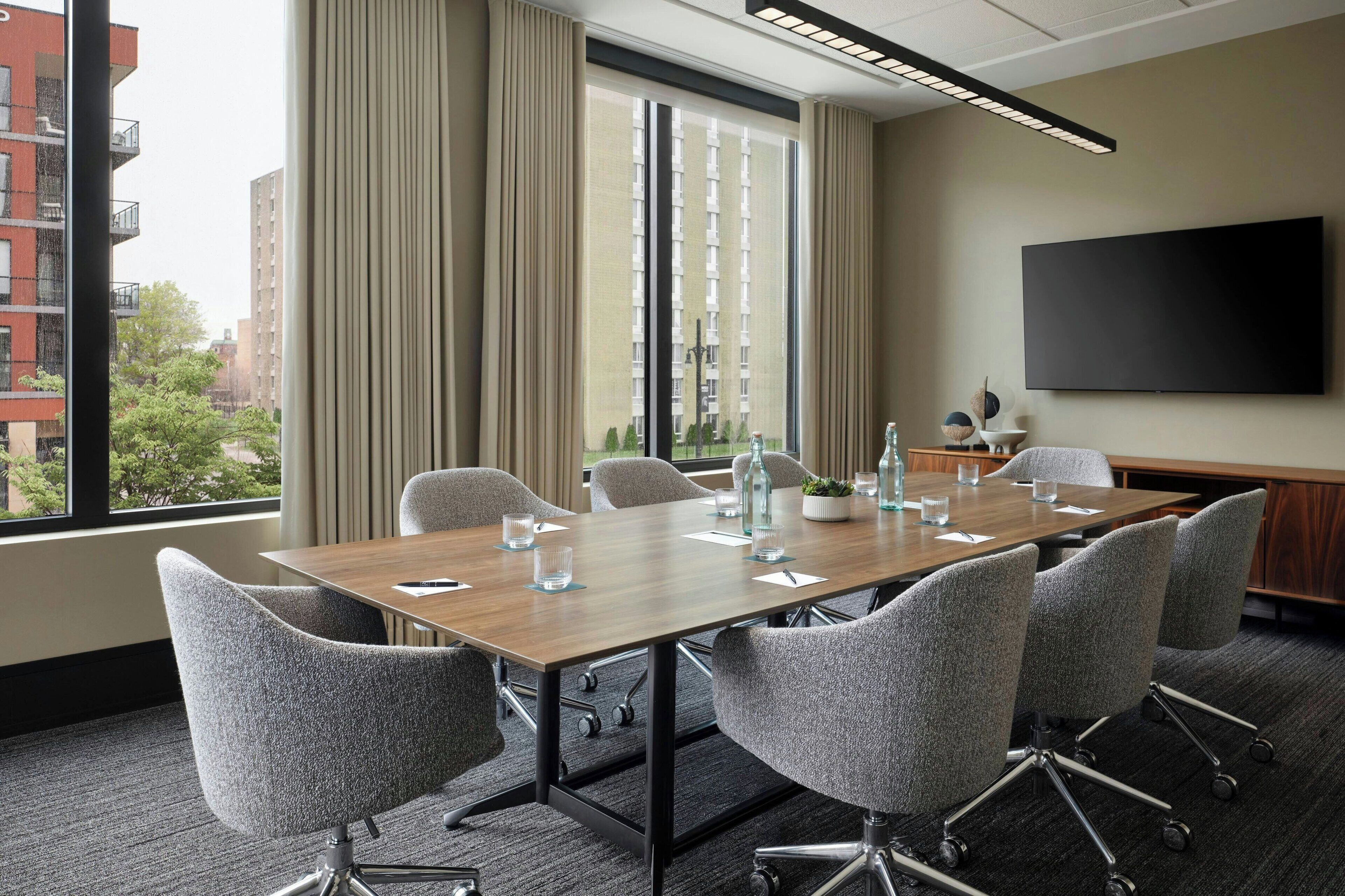 A conference room with a long table, grey chairs, a flat-screen TV, and a window with curtains.
