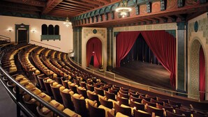 A theater with rows of wooden seats, a stage with red curtains, and ornate architectural details. - AC Hotel by Marriott Detroit at the Bonstelle (Detroit)