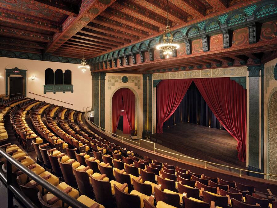 En teater med rader av trästolar, en scen med röda draperier och utsmyckade arkitektoniska detaljer.