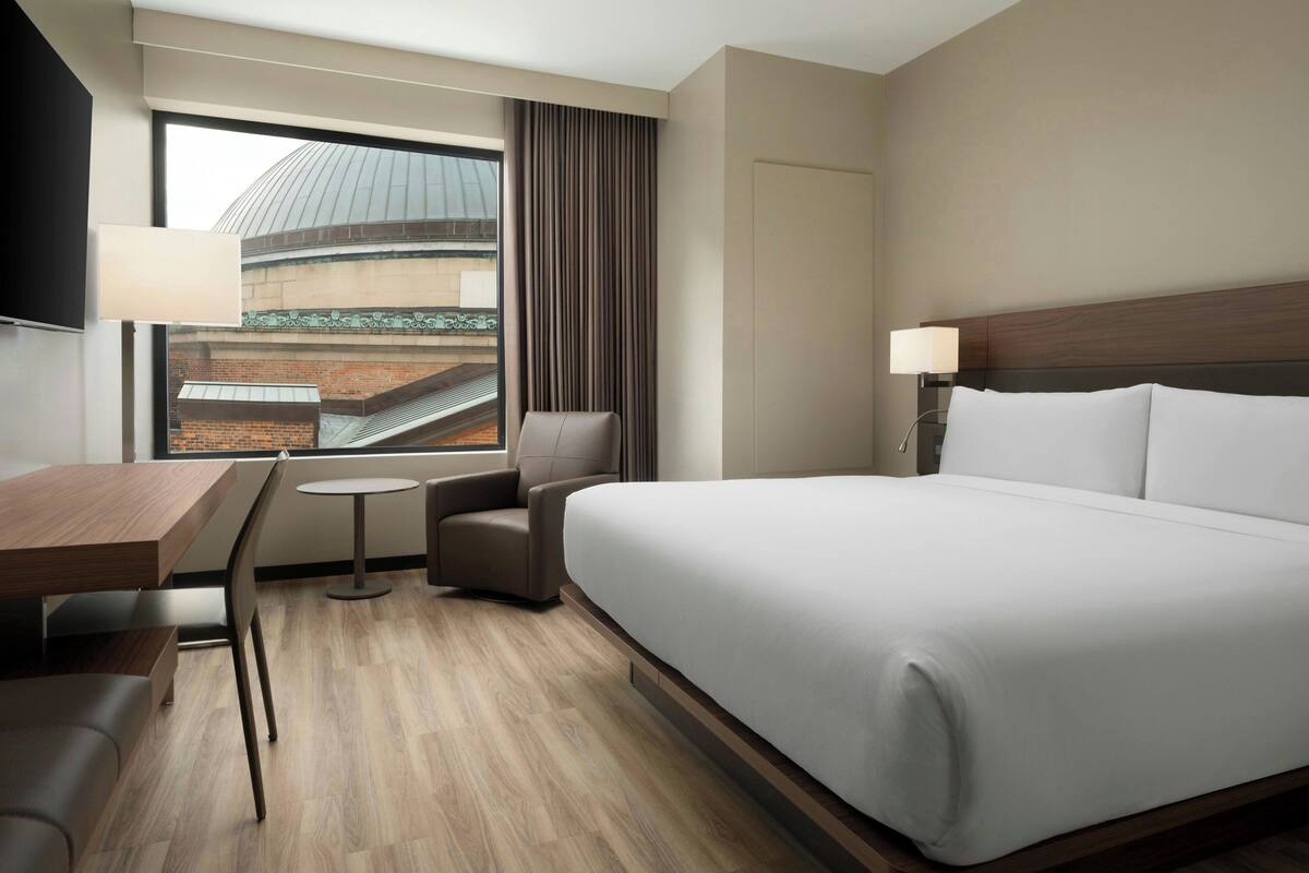 A hotel room with a large bed, a desk, a chair, a small table, and a window with a view of a building.
