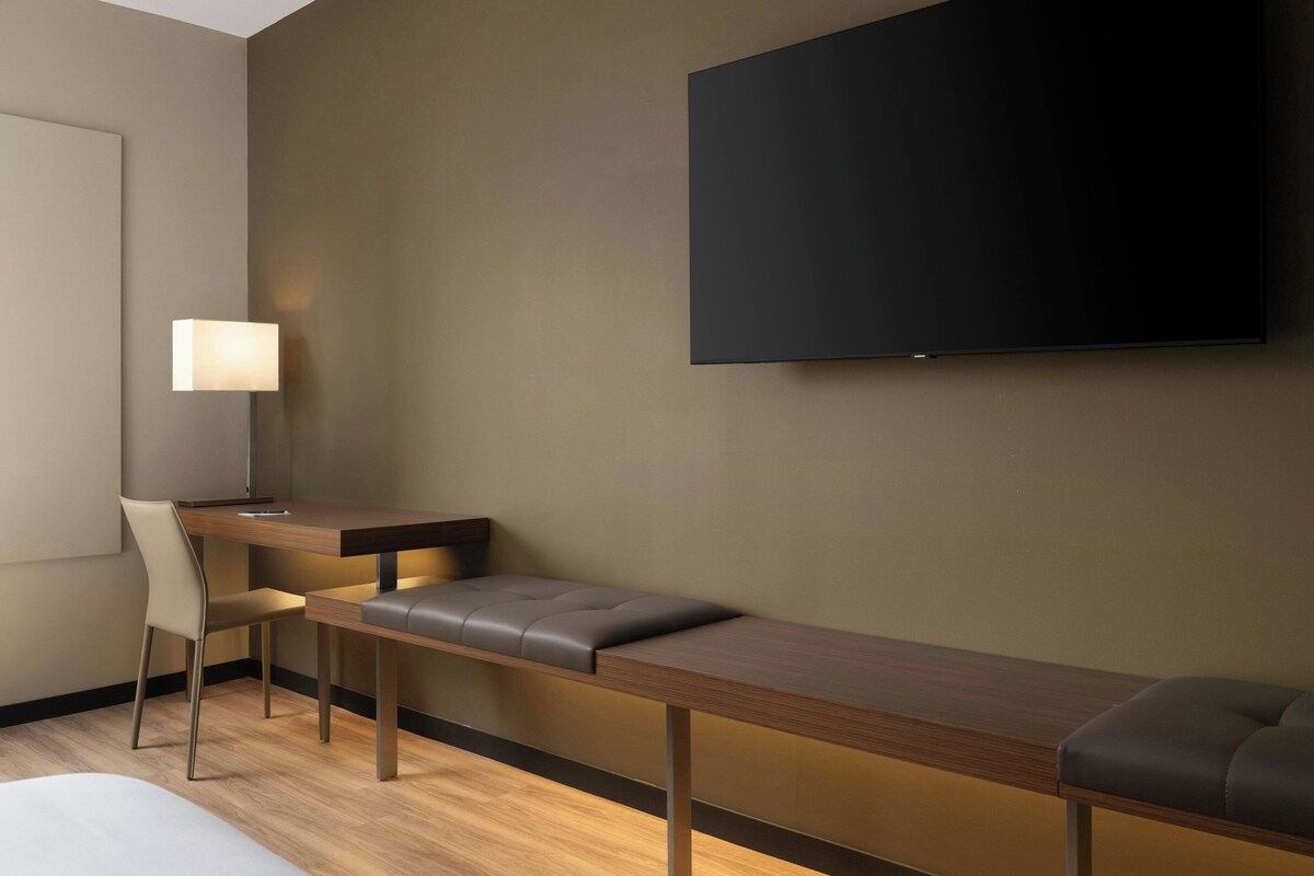 A modern hotel room with a long wooden bench, a desk, a chair, and a flat-screen TV mounted on the wall.