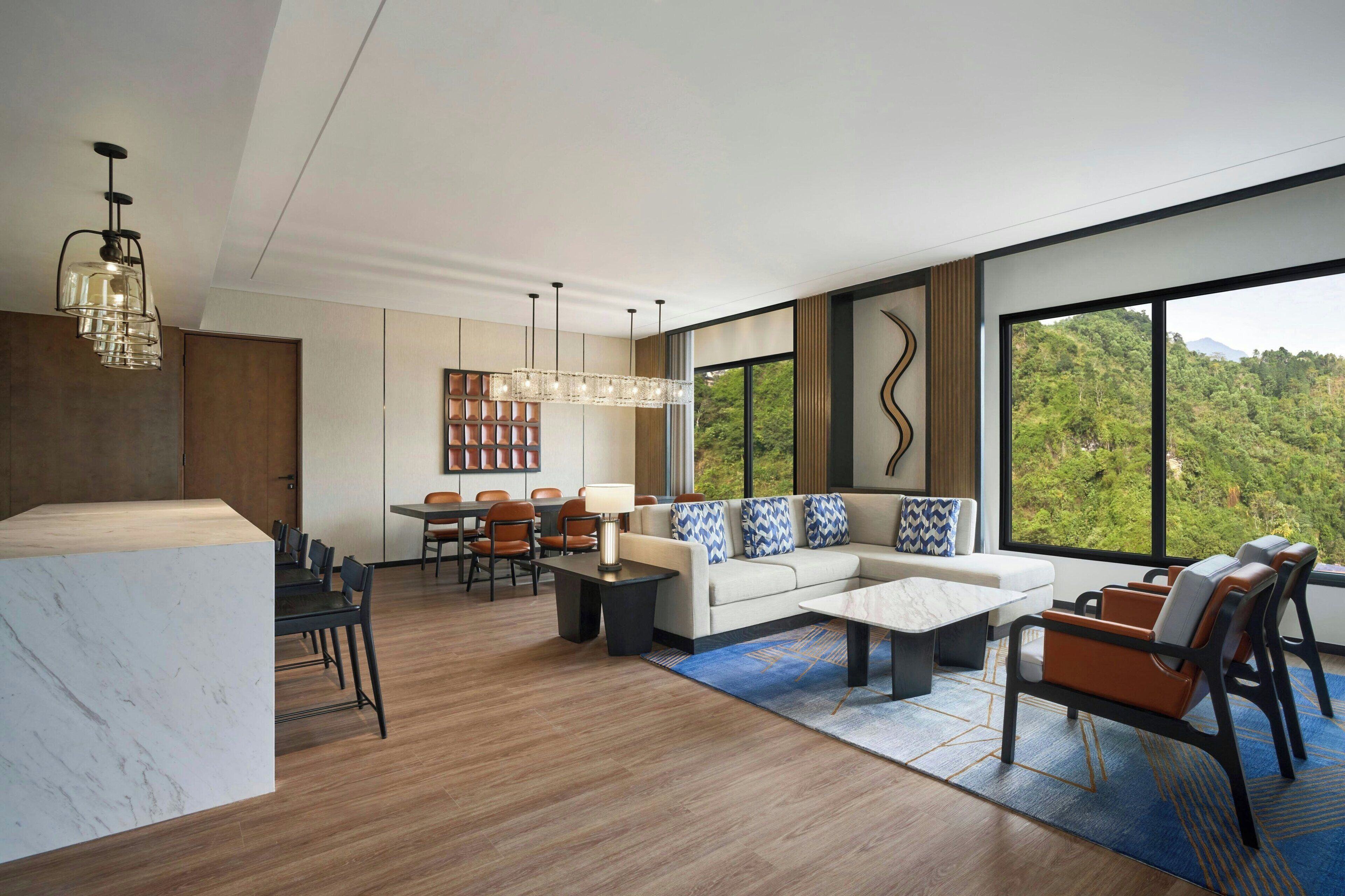 A modern hotel room with a marble reception desk, a dining area with a table and chairs, a sofa with patterned cushions, and large windows offering a view of the outdoors.