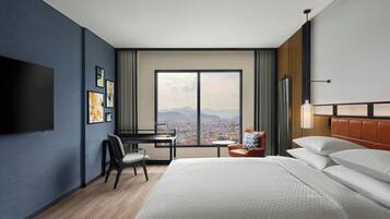 Habitación de hotel con una cama grande, un escritorio con silla, un televisor montado en la pared y vistas de un paisaje urbano a través de un ventanal amplio.