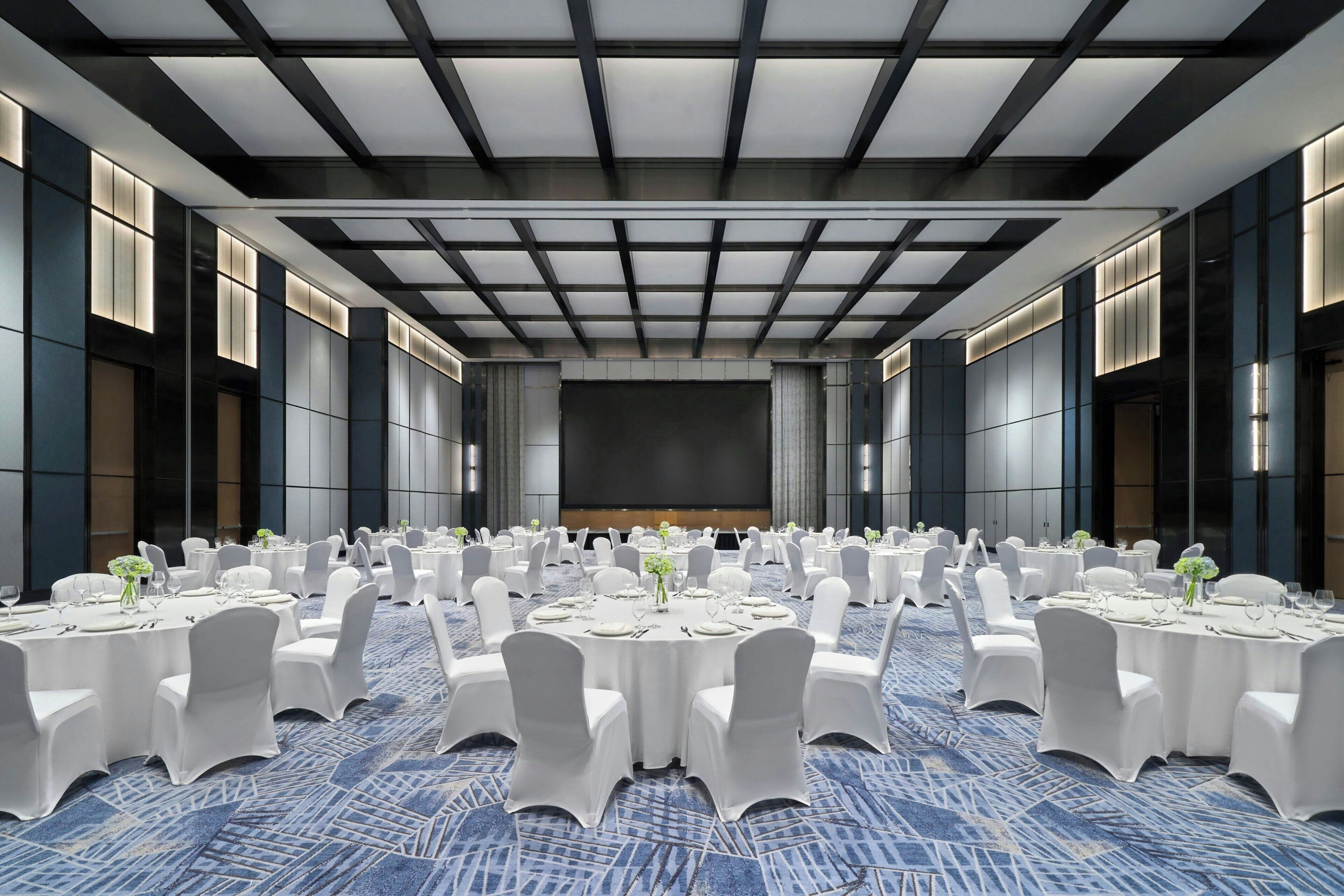 Un amplio salón de conferencias con mesas redondas, sillas blancas y alfombra azul.