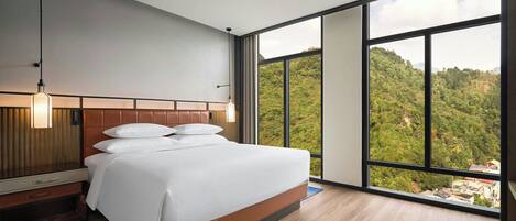 Camera d'albergo moderna con un grande letto, una testiera in legno, un comodino, una lampada e vista su un pendio boscoso.