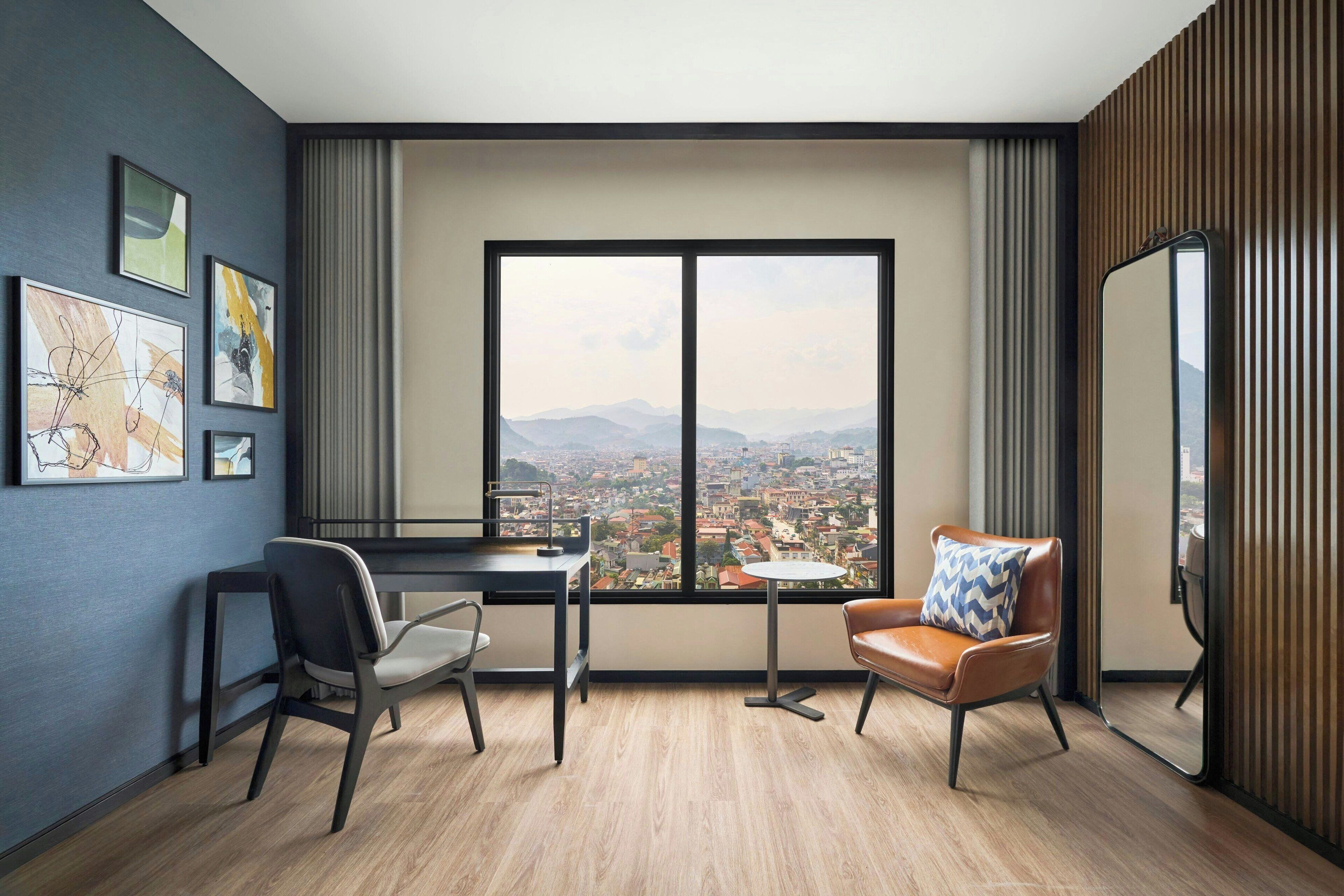Una habitación de hotel moderna con un ventanal grande que ofrece vistas a la ciudad, un escritorio con una silla, una mesita pequeña y un sofá con un cojín.