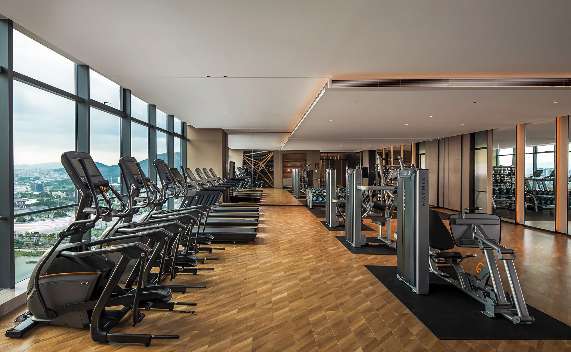 Un gimnasio con cintas de correr, máquinas de ejercicio y vistas a un paisaje urbano a través de grandes ventanales.