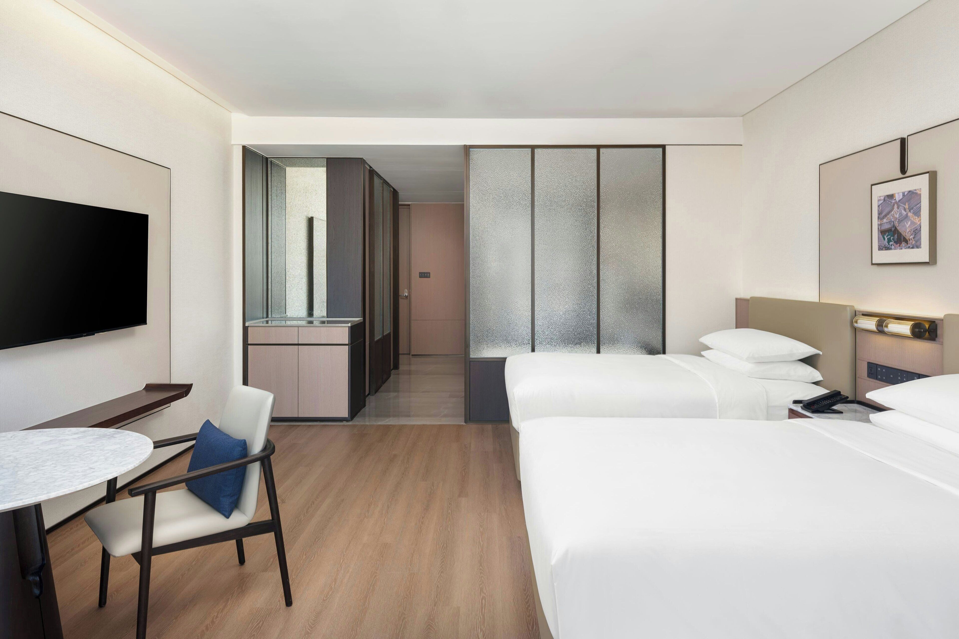 A hotel room with a bed, a desk, a chair, a TV, and a bathroom visible through a glass door.