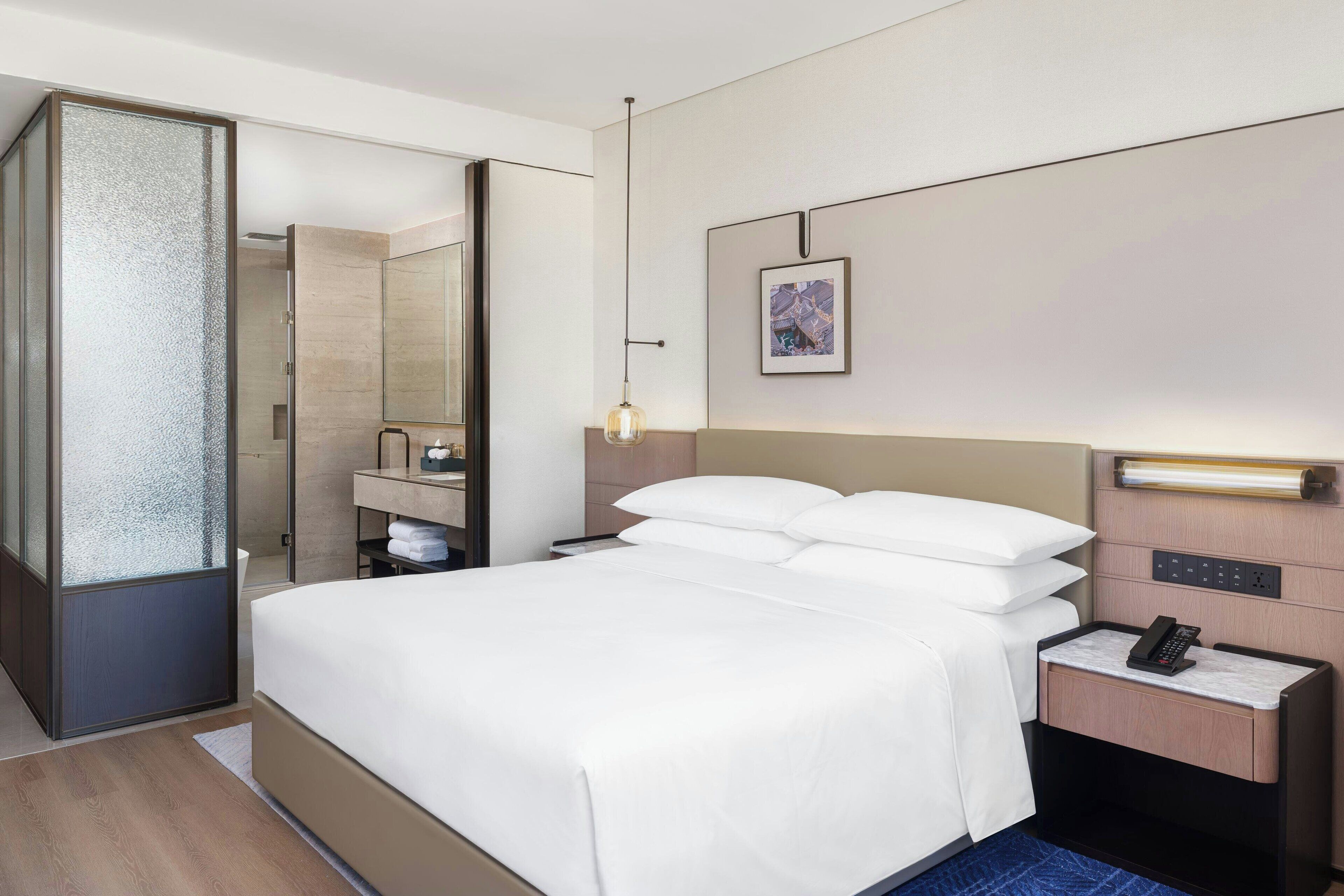 A modern hotel room with a large bed, a bedside table with a phone, a mirror, and a framed picture on the wall.