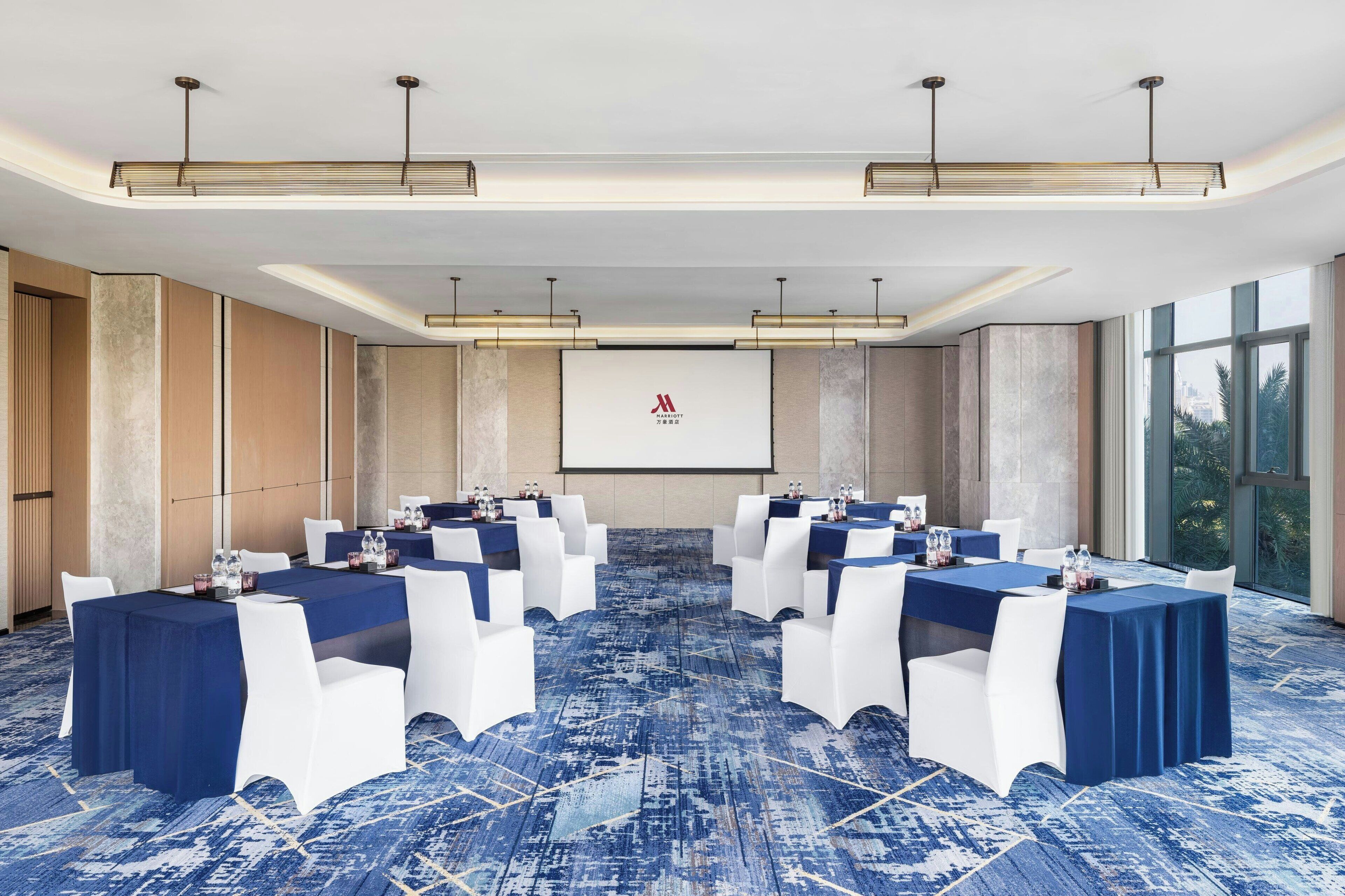 Una sala de conferencias con alfombra azul, sillas blancas y mesas dispuestas para una reunión.