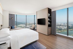 Habitación de hotel moderna con una cama grande, un televisor de pantalla plana, una estantería integrada y vistas a la ciudad a través de ventanas amplias.