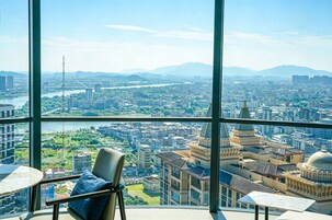 Quarto de hotel moderno em um prédio alto, com vista panorâmica da cidade, uma cadeira e uma mesa pequena.