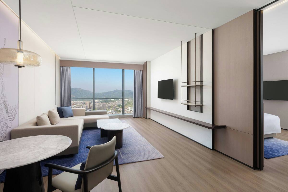 Une chambre d’hôtel moderne dotée d’une grande fenêtre, d’un téléviseur à écran plat, d’un canapé, d’une table à manger et d’un petit balcon.