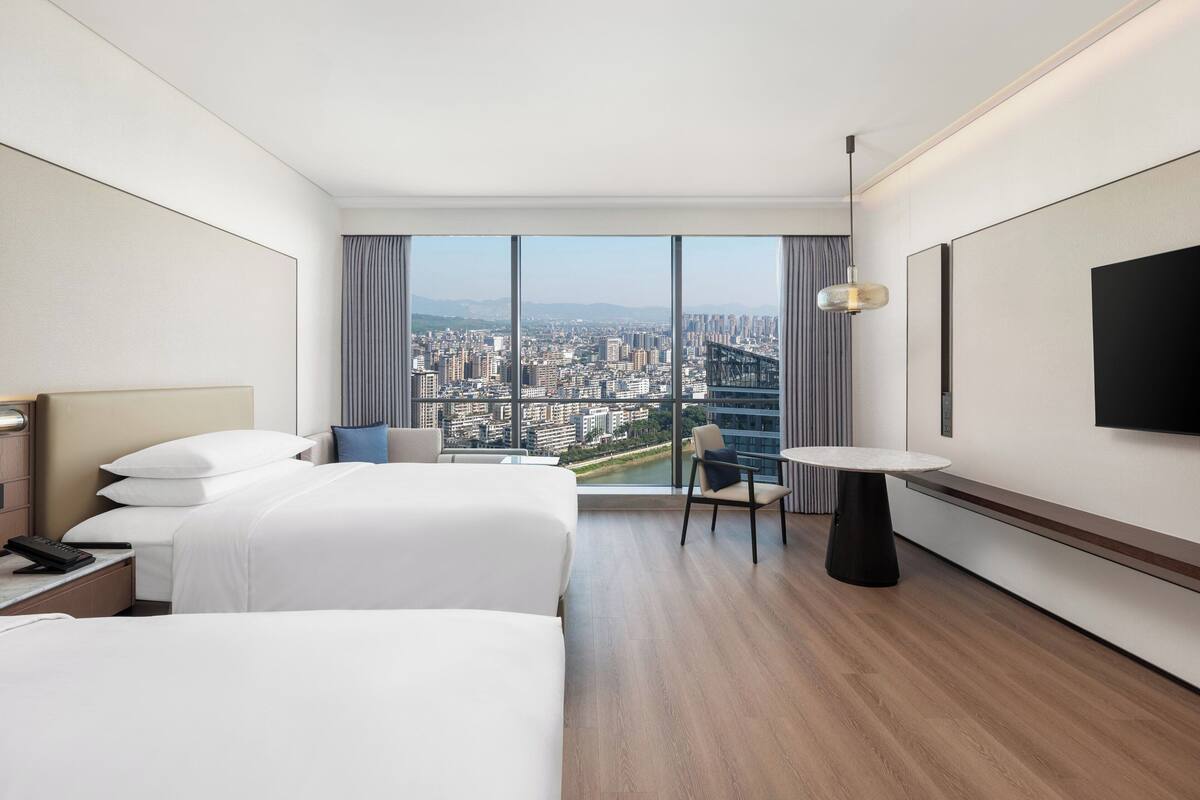 Une chambre d’hôtel moderne avec deux lits, une petite table, une chaise et une grande fenêtre donnant sur la ville.