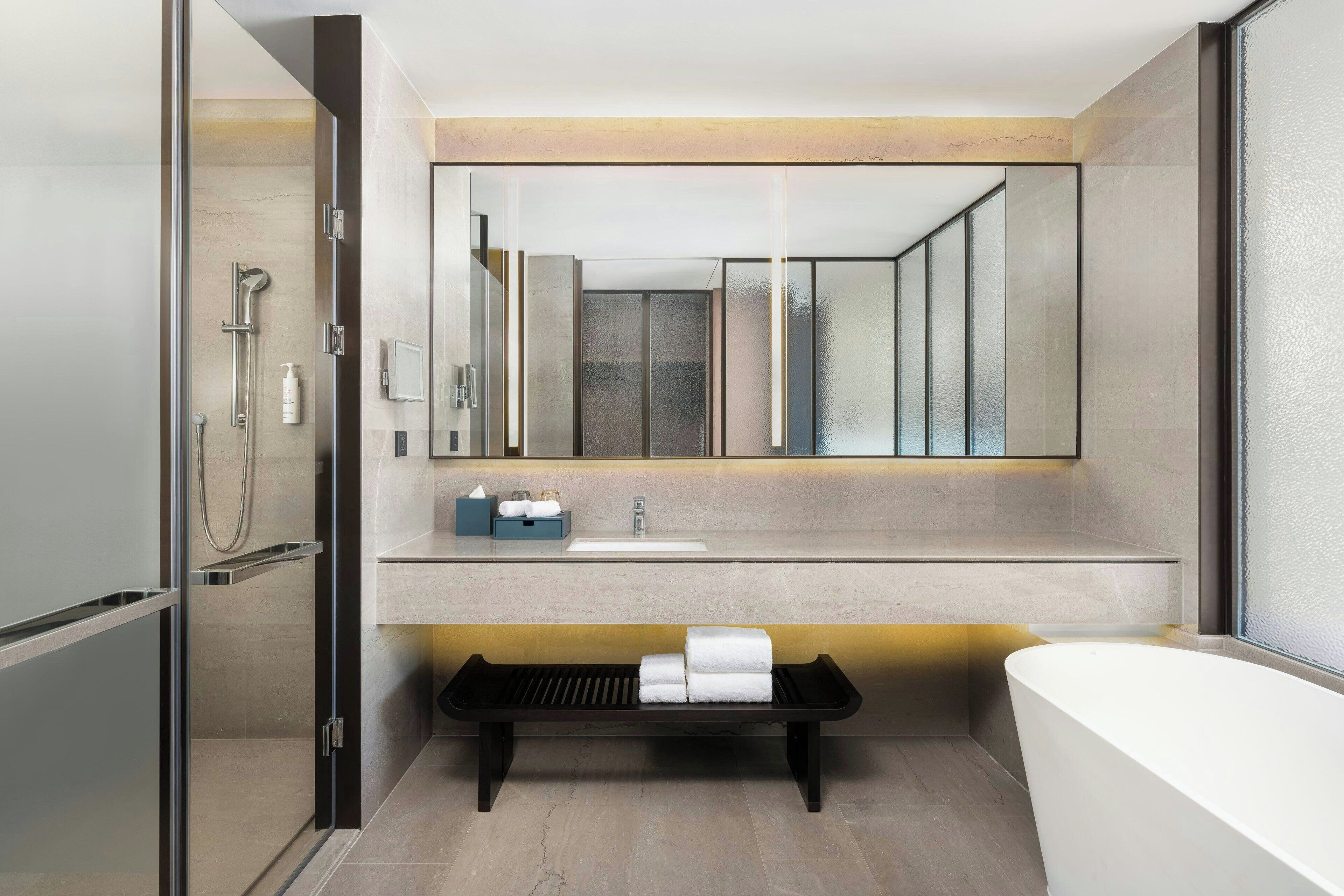 Un baño moderno con ducha de mampara de vidrio, un espejo grande, una bañera y un banco con toallas.