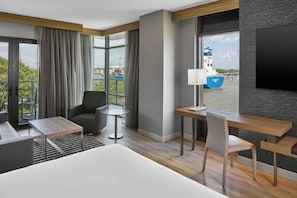 A modern hotel room with a large bed, a desk, a chair, a small table, and a view of a blue ship through large windows.