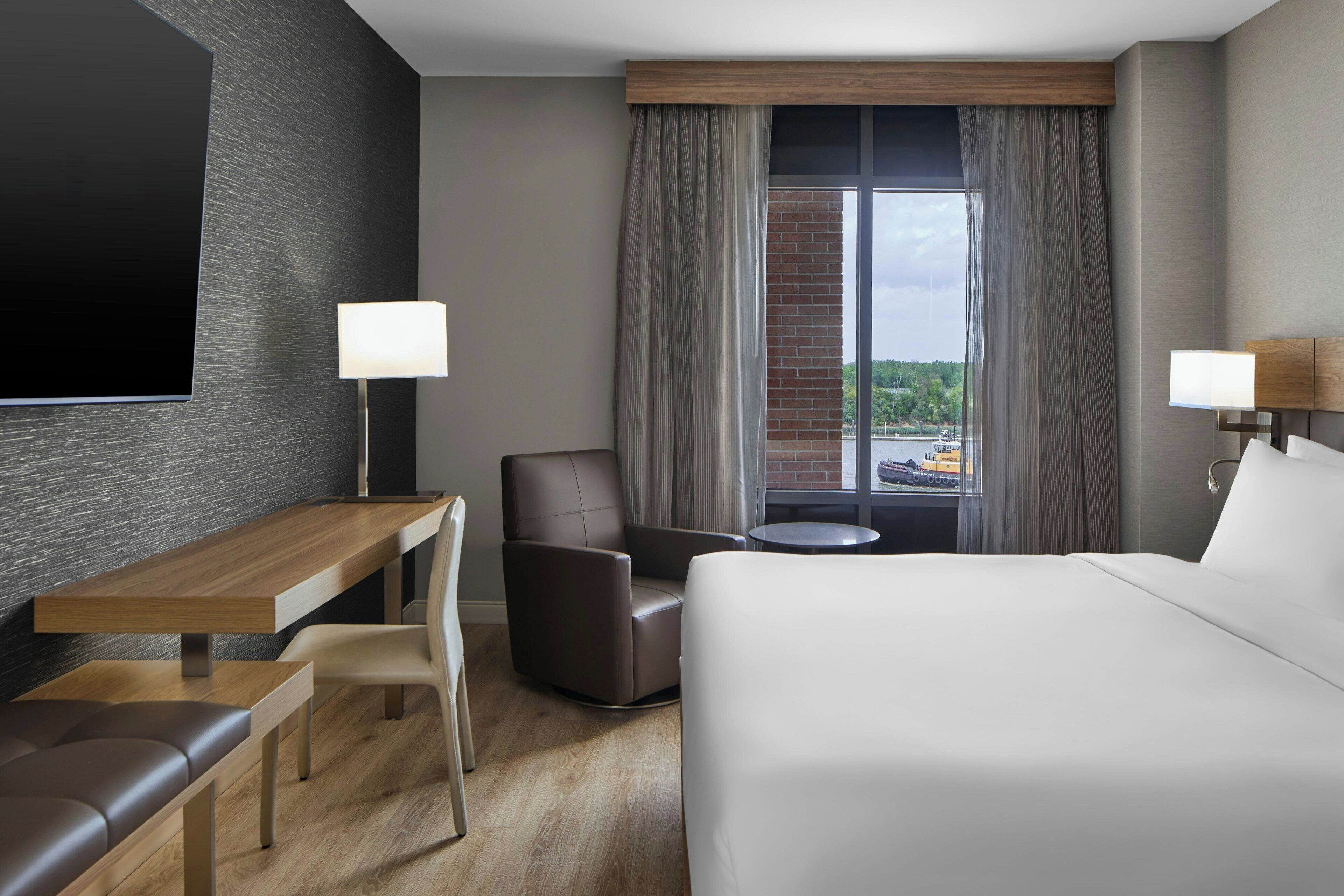 A hotel room with a bed, a desk, a chair, a small table, and a TV mounted on the wall.