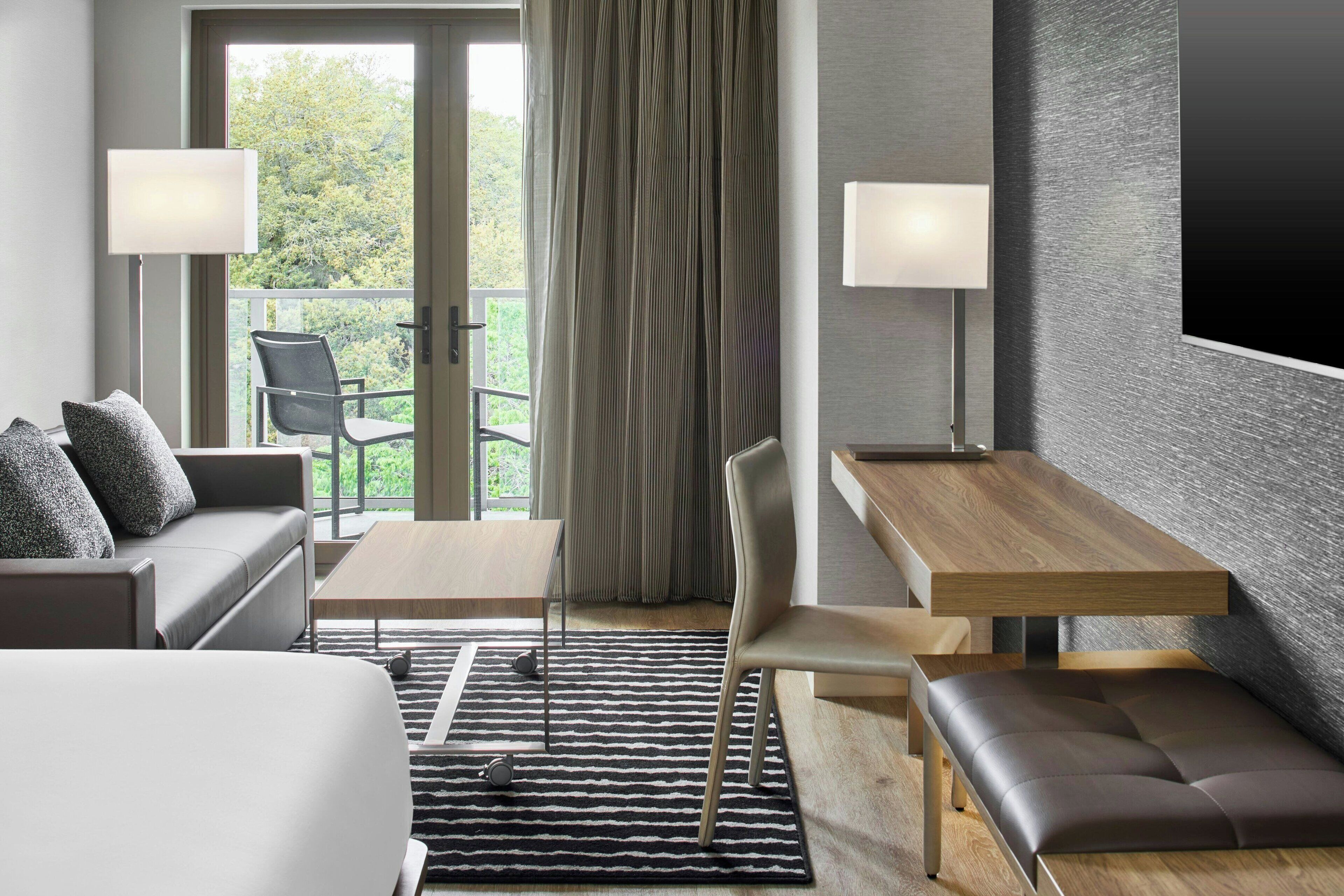 Ein modernes Hotelzimmer mit einer Couch, einem kleinen Tisch, einer Bank und Blick ins Freie durch eine Glastür.
