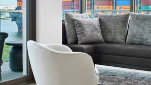 A modern living room with a white armchair, a dark sofa, and a patterned rug. Large windows offer a view of shipping containers and a bridge.