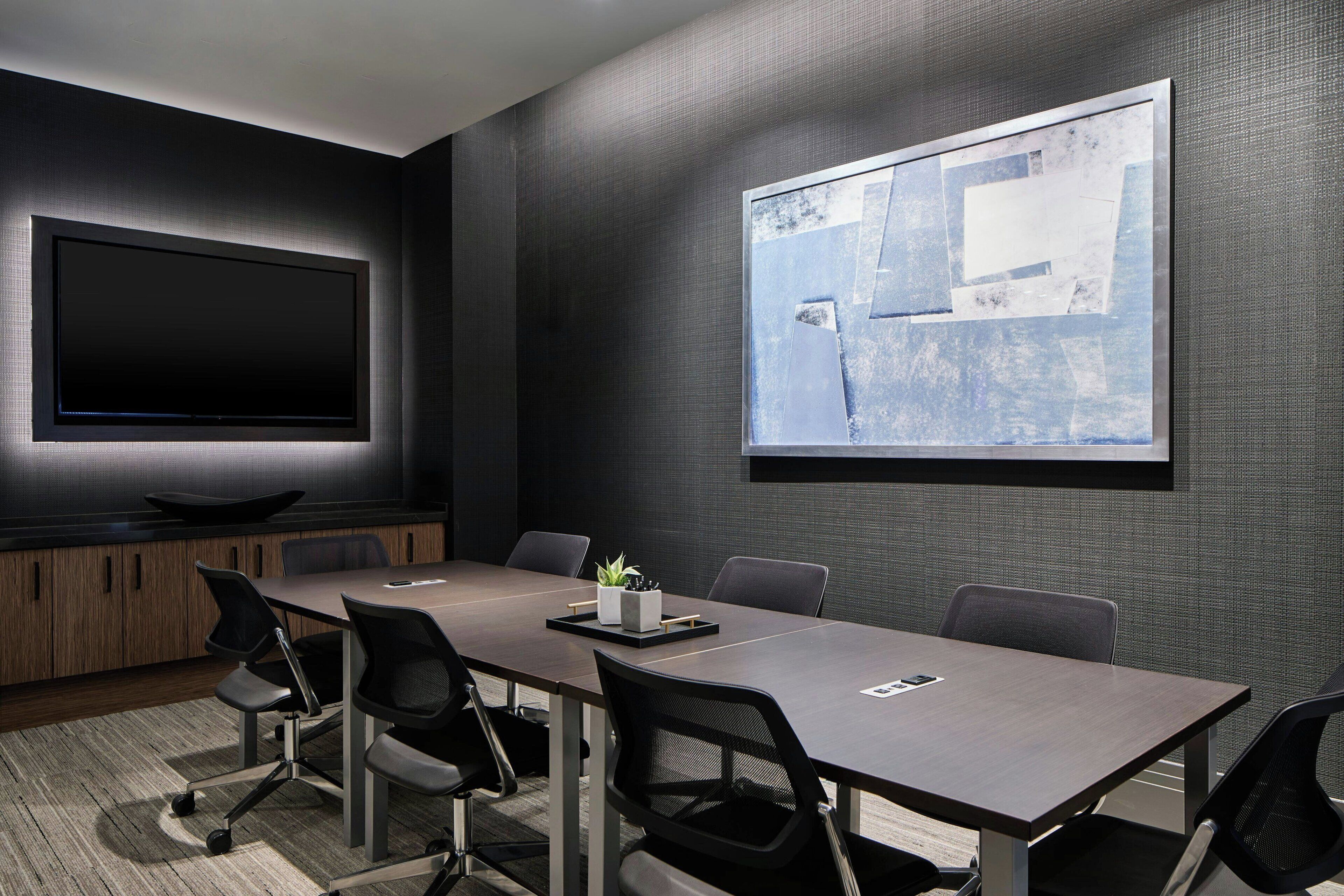 A conference room with a long table, six chairs, a flat-screen TV, and a projector screen on the wall.