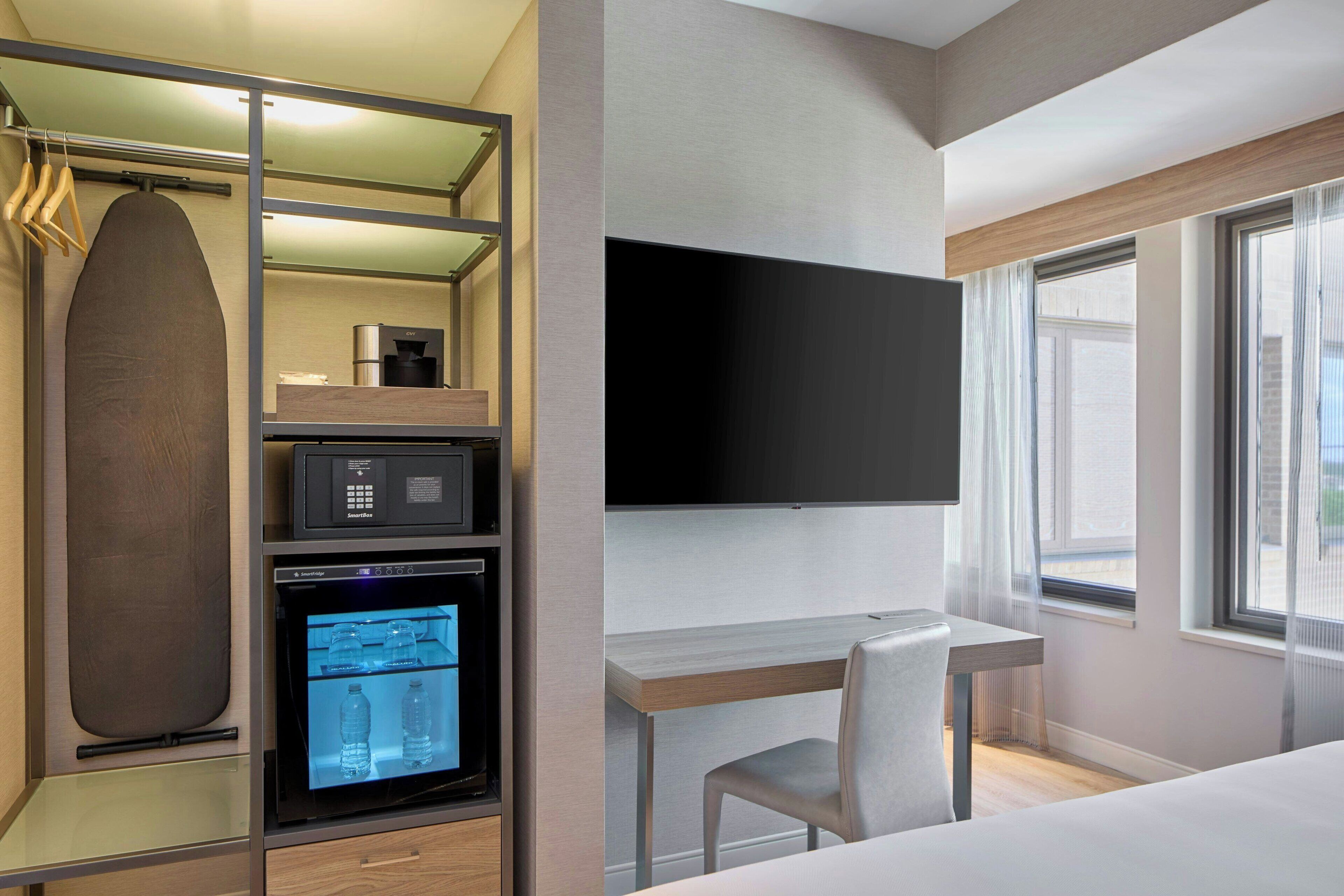 A compact hotel room with a bed, a desk, a TV, a mini-fridge, and a built-in ironing station.