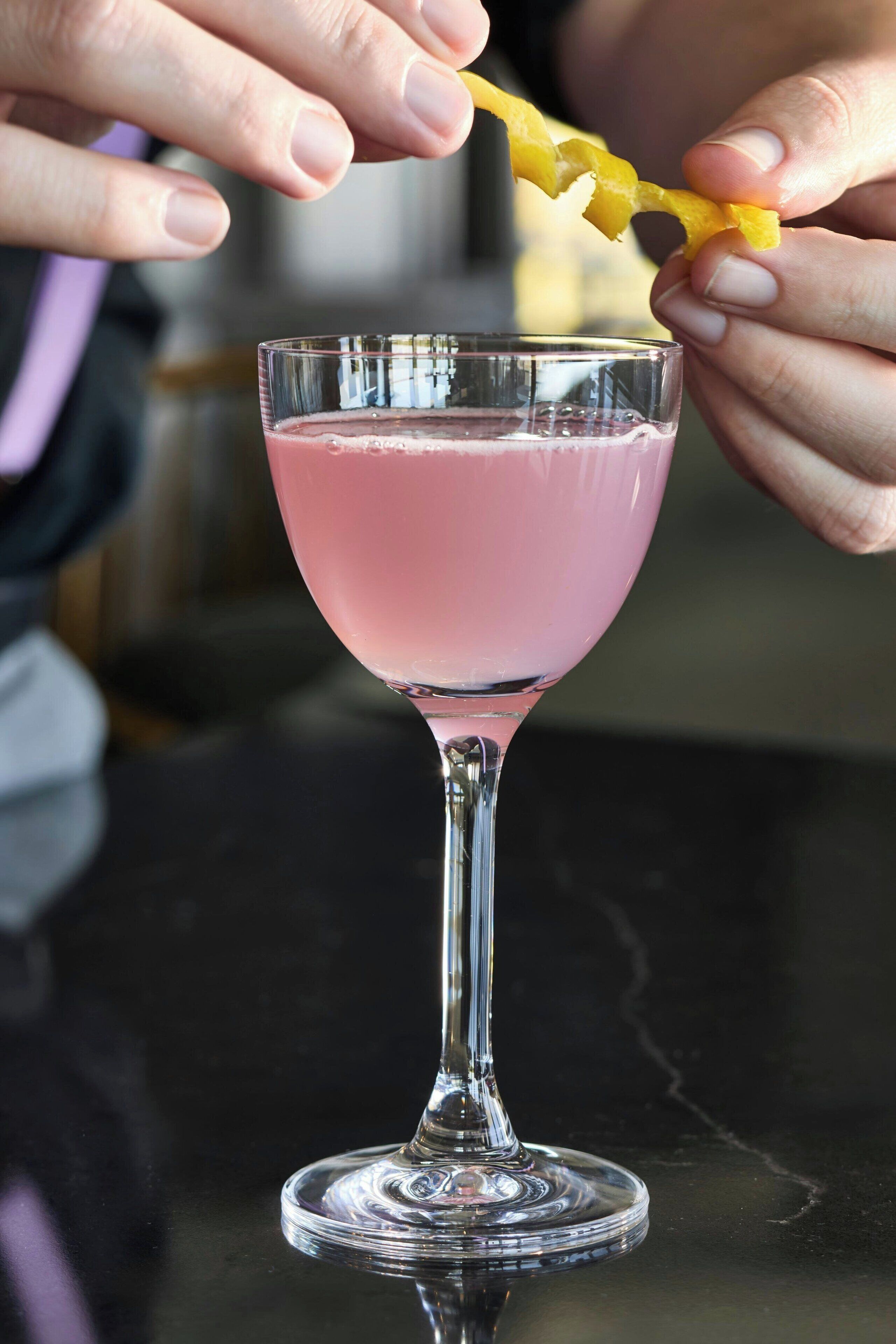 A person holding a lemon peel above a pink drink in a glass.